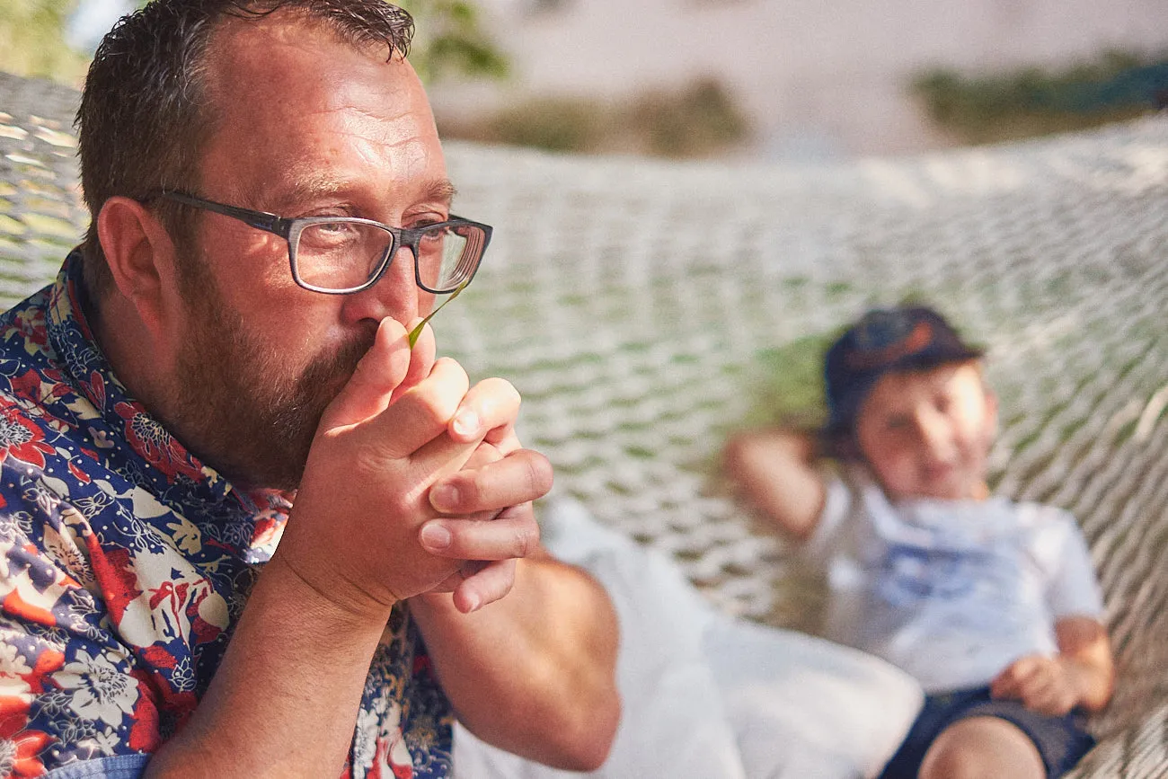 Ein Papa pustet im Vordergrund lustige Geräusche mit einem Blatt zwischen seine Händen und im Hintergrund ist sein Sohn in der Unschärfer wie er seinen Papa dabei neugierig beobachtet.