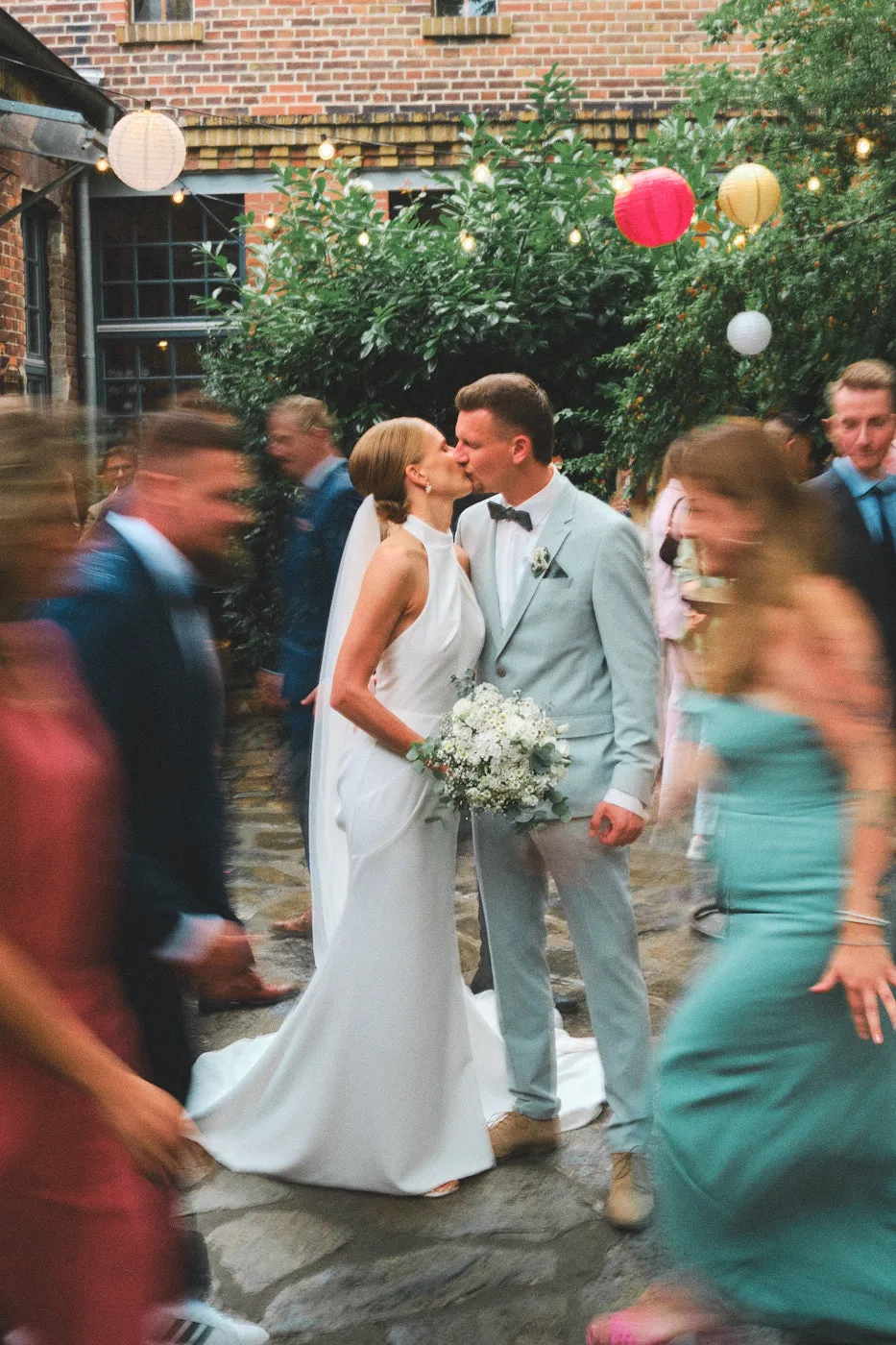 Ein Hochzeitspaar steht und küsst sich. Um sie herum bewegen sich alle ihr Gäste und sind verschwommen dargestellt.