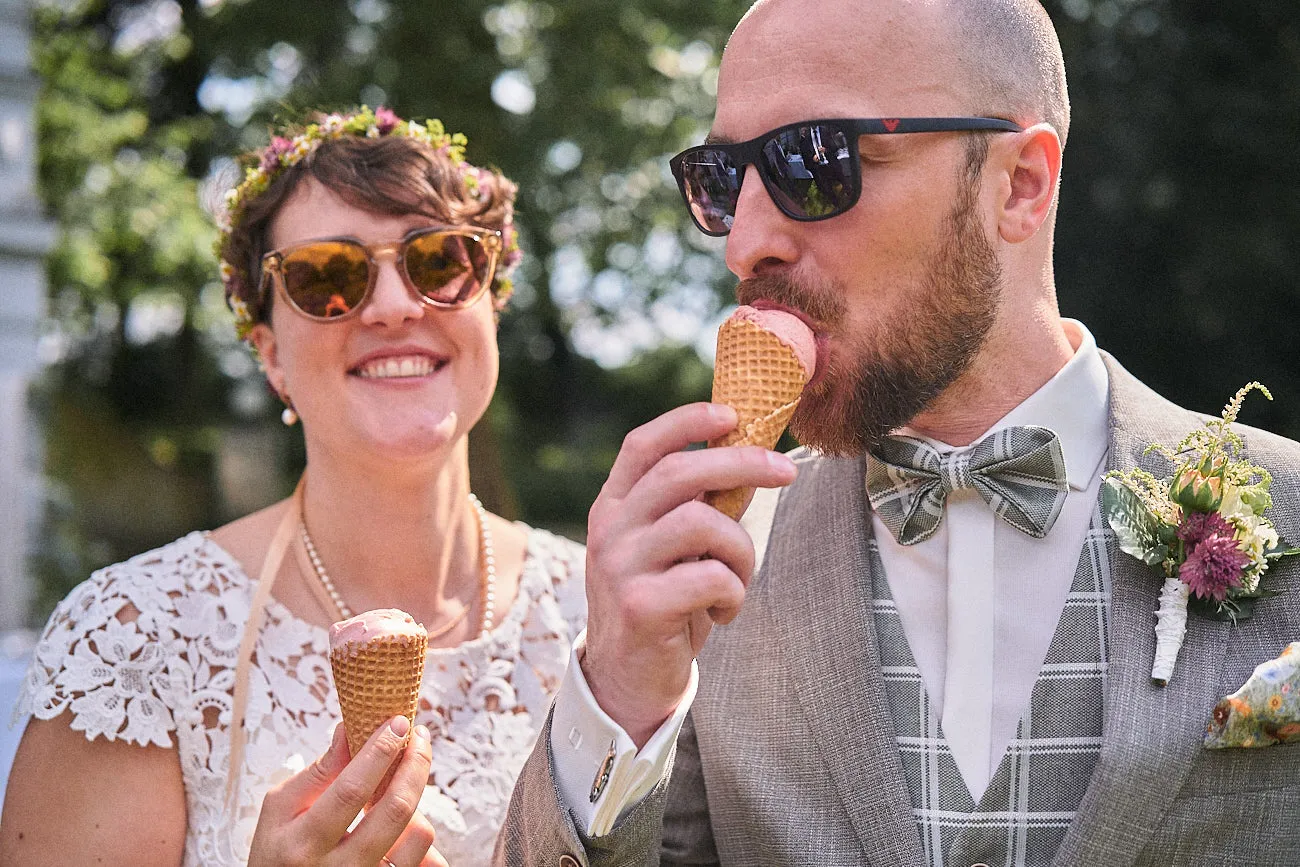 Ein Hochzeitspaar mit  Sonnenbrillen und Eis in den Händen. Die Braut lacht während der Bräutigam an seinem Eis nascht.
